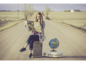 Junge Frau sitzt auf einem Koffer, der mitten auf einer langen geraden Strasse steht. Neben ihr steht ein Globus, in der Hand hat sie eine Strassenkarte. Sie macht sich gleich auf den Weg.